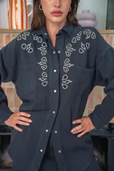Camisa negra de manga larga con cuello clásico, cierre frontal con botones a presión y dos bolsillos de parche en el pecho. Presenta apliques de strass plateados en la parte superior delantera.