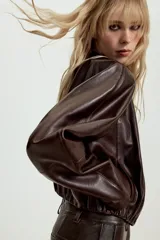 Chaqueta corta de piel color marrón, con cuello redondo, manga larga y cierre frontal con botones. Presenta un diseño con elástico en el ruedo y forro interior.