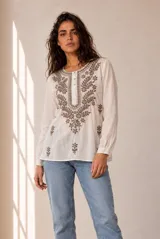 Blusa blanca de manga larga con detalles bordados en tono marrón en el pecho y las mangas, cuello redondo y cierre frontal con pequeños botones.