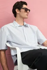 Camisa de manga corta en popelina de algodón a rayas verticales celestes y blancas. Modelo boxy con cuello inglés, tapeta clásica, bolsillo superior abierto y canesú en la espalda. Bajo recto con aberturas laterales.