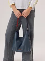 Cartera tipo hobo de cuero azul con textura granulada. Presenta un diseño holgado, con una correa de hombro bicolor que combina azul y rojo, sujeta por argollas metálicas. Incluye una placa metálica pequeña con el logo de la marca en la parte inferior frontal.
