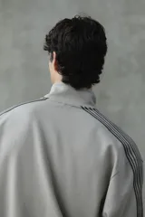 Campera deportiva de color gris topo, con cuello alto, cierre frontal bidireccional y bolsillos laterales. Presenta franjas laterales negras en las mangas y puños y bajo de punto acanalado.