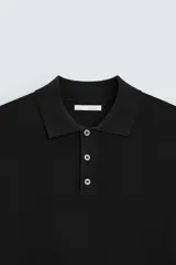 Polo de punto negro, manga corta, con cuello solapa y cierre frontal de tres botones. Presenta un calce regular y acabados en rib en mangas y ruedo.