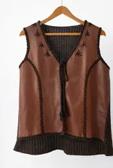 Chaleco artesanal con delantera de cuero marrón y espalda de tejido de punto marrón oscuro. Presenta detalles de bordado en lana alrededor de los bordes y costuras, escote en V y cierre frontal con cordones trenzados.