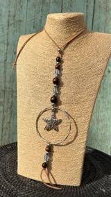 Collar largo estilo gravata con tira de cuero tostado, cuentas de madera y metal, esferas de cristal gris translúcido, un aro metálico grande y un dije de estrella de mar.