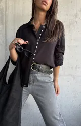 Camisa de color negro con cuello clásico y cierre frontal de botones blancos. Presenta mangas largas con detalles bordados en los puños.
