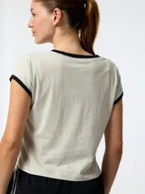 Remera de manga corta estilo crop, color gris claro con detalles en negro en el cuello y en los bordes de las mangas. Presenta un pequeño logo bordado en el pecho.
