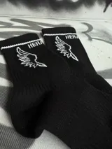 Par de medias blancas de algodón con puño acanalado con una línea negra, logo de la marca en el tobillo y estampado de alas negras.