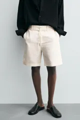 Bermuda de corte relajado color beige claro, confeccionada en tejido de algodón y lino. Presenta cintura con cordón ajustable del mismo tejido, bolsillos delanteros y bolsillos traseros de vivo. Cierre frontal con cremallera y botón.