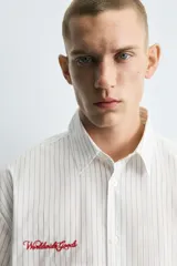 Camisa de algodón blanca con finas rayas verticales negras, corte relajado (relaxed fit) y manga corta. Presenta un bordado en hilo rojo en el lado izquierdo del pecho.