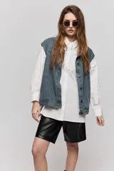 Chaleco de jean azul con sutil textura a rayas, cuello camisero, cierre frontal con botones metálicos y diseño sin mangas con hombros amplios.