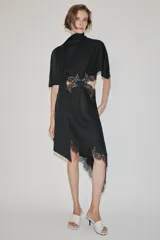 Vestido midi de satén negro con cuello subido, manga corta tipo capa y pañuelo incorporado en el mismo tejido. Presenta aberturas laterales decoradas con encaje a tono y bajo asimétrico con detalles de encaje. Cierre en la espalda con abertura y botones forrados.
