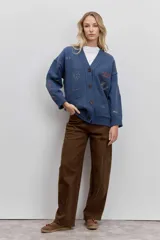 Cardigan de punto azul con cuello en V, cierre frontal de botones y bolsillos delanteros. Presenta bordados decorativos con motivos variados y frases en colores contrastantes.