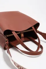 Bolso de hombro de tamaño mediano color marrón, confeccionado en piel vacuna. Presenta doble asa corta de mano y correa de tejido jaspeada con el nombre de la marca estampado. Incluye detalles en piel, cierre con botón de presión, logo grabado en dorado en el frente y acabados metálicos.