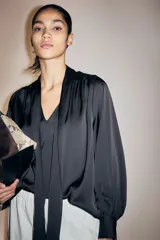 Blusa de gasa negra con cuello alto y lazada frontal, mangas largas abullonadas con puños abotonados y detalle de frunces en los hombros.