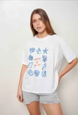 Remerón de algodón color blanco, de corte oversize, manga corta y cuello redondo. Presenta un estampado central con ilustraciones de caracoles y conchas marinas en azul, junto con el texto "Meet me by the sea" en tono rojizo.