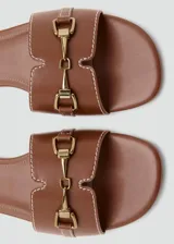 Sandalias planas de color marrón, con tira ancha en el empeine decorada con costuras blancas y un detalle metálico dorado. Diseño destalonado y puntera redondeada.