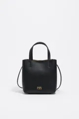 Bolso pequeño tipo tote o cubo de piel vacuna texturizada color negro, con asas cortas superiores y correa larga ajustable para llevar al hombro o cruzado. Presenta el logo metálico de Bimba y Lola en dorado en el frente.