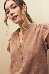Camisa color beige con textura suave, cuello mao con volado y mangas cortas con pequeño volado. Presenta botonadura frontal.