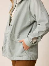 Campera de corte recto y estilo funcional, color beige, con capucha integrada, cierre frontal con botones a presión y bolsillos delanteros amplios. Presenta un diseño minimalista con cordones ajustables en la capucha.