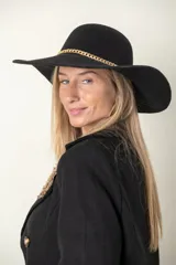 Sombrero tipo capelina de paño color negro, con ala ancha y detalle de cadena dorada en la base de la copa.