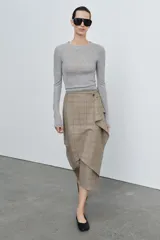 Falda midi de tiro medio con estampado de cuadros en tonos beige y gris. Presenta un diseño cruzado con un volante decorativo en el delantero y cierre con botón.