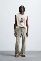 Camiseta tank de punto color beige claro, corte relajado (relaxed fit) y cuello redondo. Presenta un estampado central de un vehículo todoterreno cargado de equipaje conduciendo por una carretera desértica. Los bordes del cuello y las sisas tienen un acabado deshilachado.