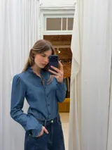 Camisa de denim fino en tono azul medio, con cuello alto con volados, mangas largas con puños con volados y cierre frontal con botones en contraste color rojo.