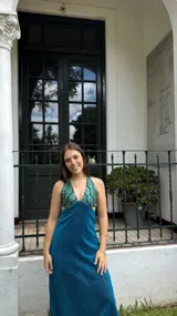 Vestido largo con estampado tie-dye en tonos violeta y fucsia, escote en V con detalles bordados dorados, cut-outs laterales bajo el busto y tirantes finos que se cruzan y atan en la espalda.