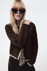 Chaqueta de punto de textura bouclé en color marrón oscuro, con cuello redondo y manga larga. Presenta detalles de vivo a contraste en color crema en los bordes y cierre frontal mediante lazadas decorativas con flecos.
