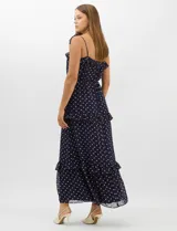 Vestido largo azul marino con estampado de lunares blancos, confeccionado en gasa fluida. Cuenta con tirantes finos ajustables con lazos en los hombros, escote con volados, corte en la cintura con lazo ajustable, y falda larga con múltiples capas y volados horizontales.