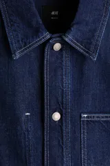 Sobrecamisa de denim de algodón rígido, color azul, con cuello inglés, bolsillos de parche en el pecho y en la parte inferior, y cierre frontal de botones metálicos.