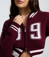 Cardigan de punto estilo college color bordó, con cierre frontal de doble cremallera, mangas largas y detalles de rayas blancas en cuello, puños y ruedo. Presenta una estampa numérica blanca en el frente.
