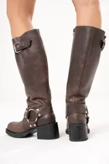 Bota biker de caña alta color marrón, confeccionada en cuero. Presenta hebilla en la parte superior, detalle de aro en la parte inferior y cierre lateral.