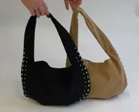Cartera tipo hobo de gamuza color marrón claro, con detalles de tachas plateadas en los bordes y las asas. Cuenta con bolsillo interno con cierre.
