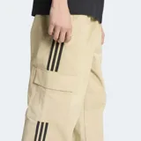 Pantalón cargo Adidas de corte holgado, color beige, confeccionado en tejido antidesgarre. Presenta las icónicas tres rayas laterales en negro, bolsillos cargo en los laterales y logo del trifolio bordado en la pierna izquierda.