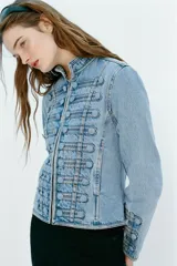 Chaqueta denim entallada de cuello redondo y manga larga, con cierre frontal de cremallera metálica. Presenta detalles de aplicaciones en el mismo tejido y pasamanería frontal de inspiración militar.