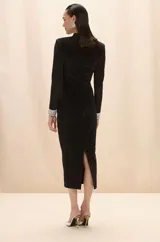 Vestido midi negro de corte ajustado, con mangas largas, cuello alto y aberturas estratégicas con paneles de tul transparente en el pecho y la cintura. Presenta puños decorados con aplicaciones de cristales brillantes.