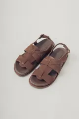 Sandalias planas de gamuza color marrón, con diseño de tiras cruzadas en el empeine y cierre con hebilla metálica dorada en el tobillo. Cuenta con plantilla de cuero y suela de goma con textura.