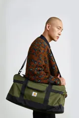 Bolso tipo duffle Carhartt WIP modelo Jack Duffle Bag, en color verde militar con refuerzos y asas negras, correa ajustable para el hombro y el parche con el logotipo de la marca al frente.