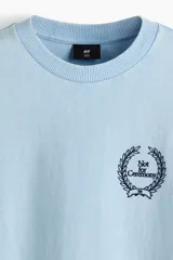 Remera de algodón color celeste, con cuello redondo, mangas cortas y corte holgado. Presenta un pequeño estampado frontal en el pecho con diseño de corona de laureles y texto.