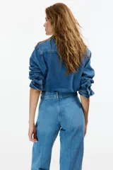 Jean de tiro alto en denim de algodón rígido, con diseño de pierna ancha y corte holgado desde el muslo hasta el bajo. Presenta cierre frontal con doble botón, bolsillos laterales y pliegues marcados en la parte delantera y trasera.