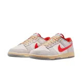 Championes Nike Dunk Low con diseño de paneles en tonos gris claro y blanco, destacando el swoosh lateral y detalles en el talón y cuello en color rojo vibrante. Presenta cordones en tono crema y una suela con efecto envejecido.