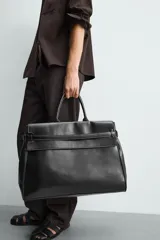 Bolso de mano extragrande negro de cuero sintético, con compartimento principal con cierre de imán, bolsillo interior con cierre y bolsillo secundario con solapa y detalle de cinta. Cuenta con dos asas de mano.