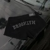 Short de denim negro con diseño de tiro bajo, bolsillos traseros con apliques de strass formando la inscripción "BROOKLYN" y terminaciones deshilachadas.