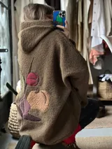 Campera tipo hoodie confeccionada en tela sherpa color verde oliva, con diseño de apliques bordados en la espalda que representan una flor y una fruta. Presenta capucha integrada y corte holgado.