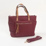 Cartera tipo bolso de color bordó, confeccionada en material textil con detalles en cuero sintético color suela. Cuenta con doble asa de mano, un bolsillo frontal con cierre y tiradores decorativos, además de una correa larga ajustable y extraíble.