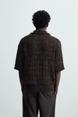 Polo de punto de calce relajado, color marrón oscuro jaspeado con negro. Presenta cuello solapa con abertura frontal en V y mangas cortas.