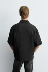 Camisa negra de manga corta con cuello solapa y textura arrugada o plisada. Corte relajado (relaxed fit) con aberturas laterales y cierre frontal de botones.