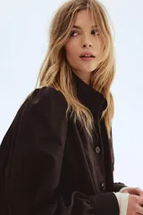 Campera corta de sarga color marrón oscuro, con cuello alto, cierre frontal con botones y bolsillos laterales. Presenta mangas largas abullonadas con fruncidos en los hombros y puños ajustados.
